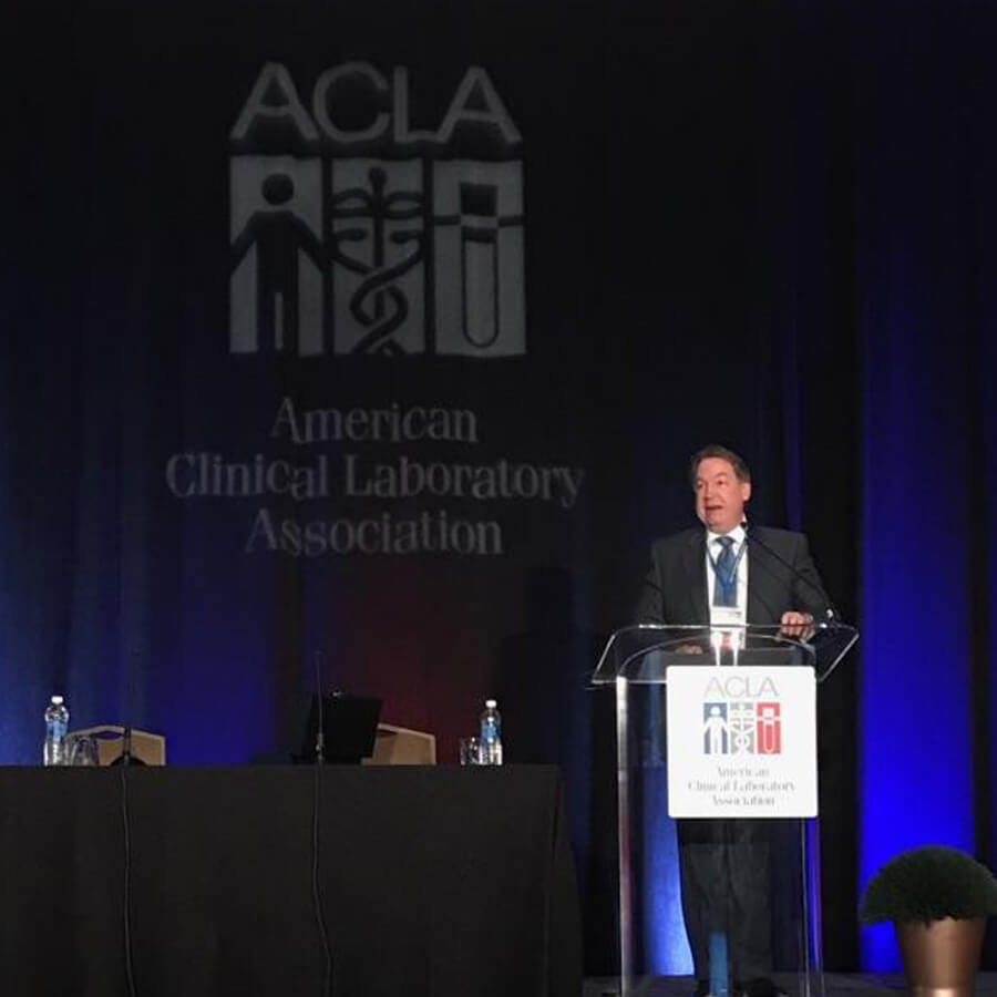 Standing at a clear podium at the front of a dark blue stage, Steve Rusckowski, Quest Diagnostics Chairman, President and CEO, addresses the American Clinical Laboratory Association (ACLA) 2017 annual meeting in Washington, D.C.